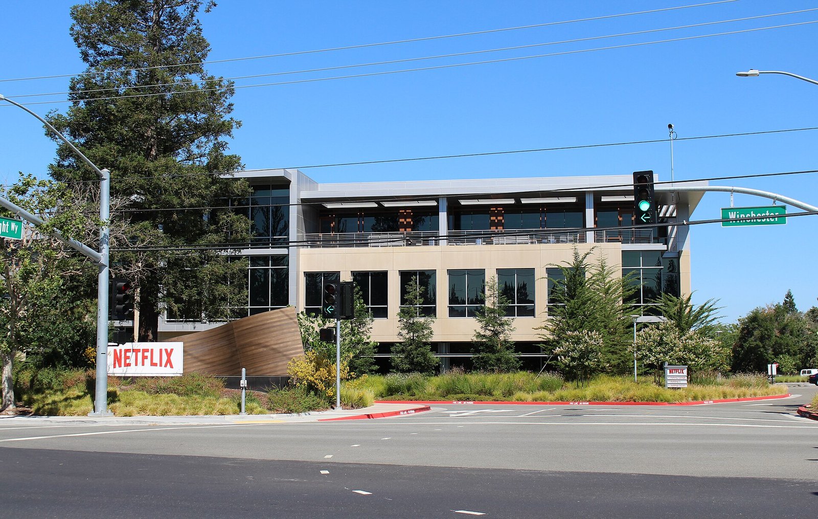 A modern three-storey office building with large windows behind trees and landscaping on a street corner, with a red Netflix sign on a low wall in the foreground under a clear sky.