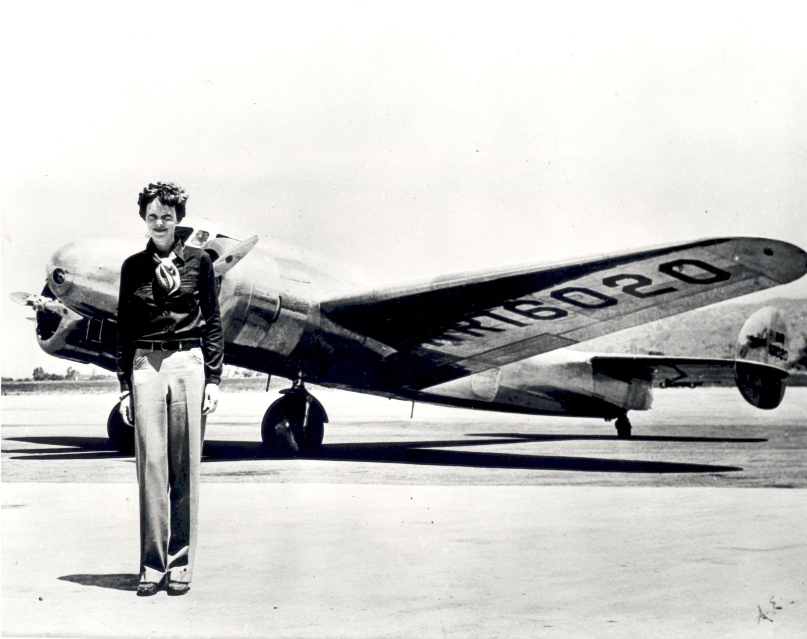 Black-and-white photo of Amelia Earhart standing in front of her silver twin-engine Lockheed Electra, registration NR16020 painted on the wing.