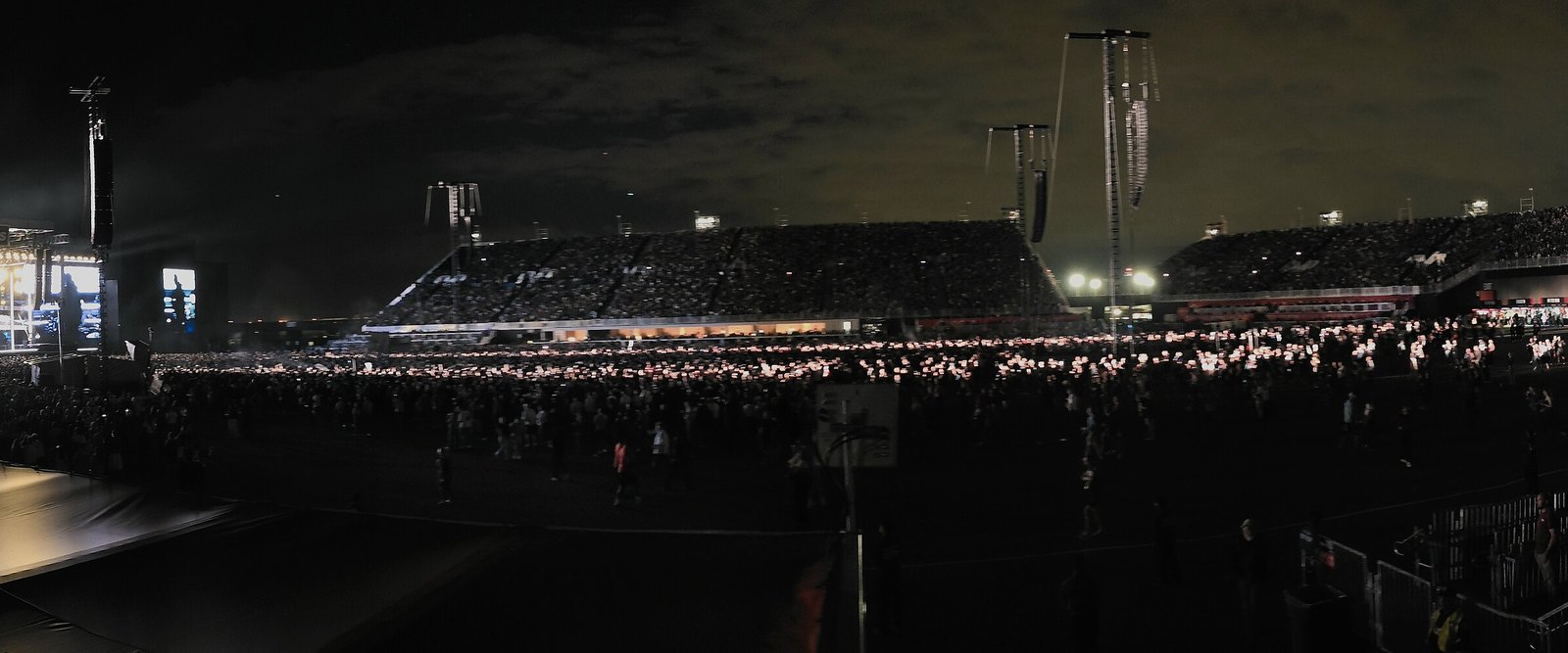 A night-time stadium crowd stretches across a vast open-air field toward a temporary grandstand, lit by thousands of phone lights.