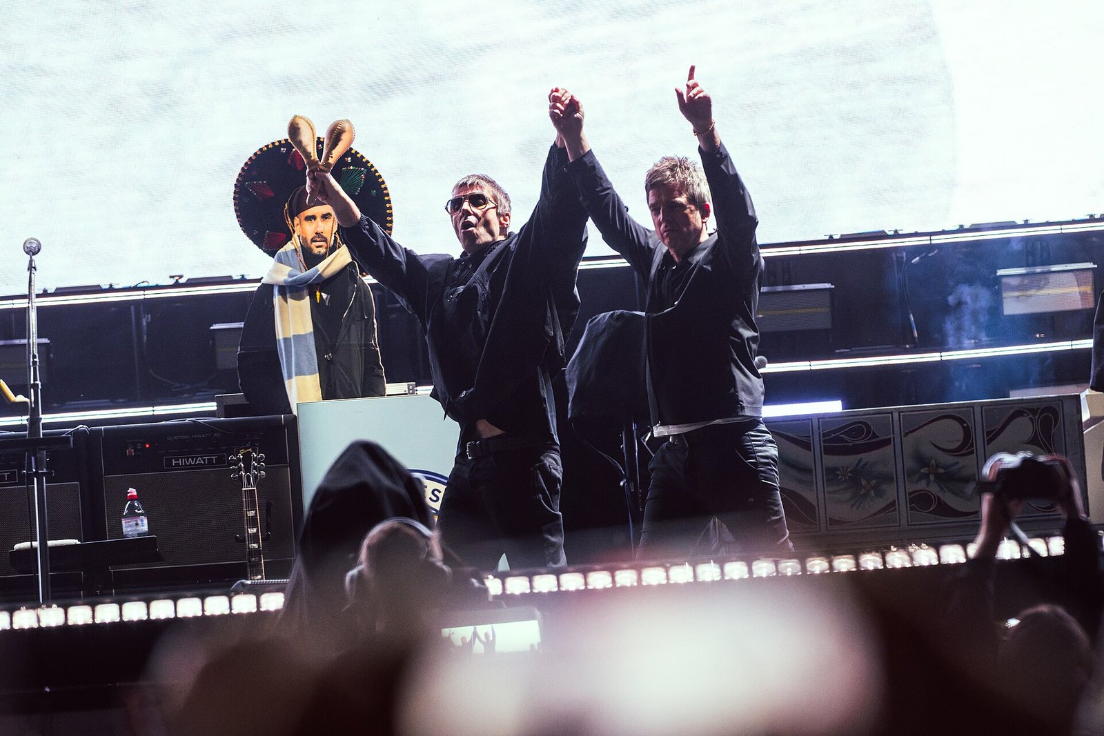 Liam and Noel Gallagher, dressed in black, raise their joined hands on a stadium stage as phone lights glow from the crowd in front of them.