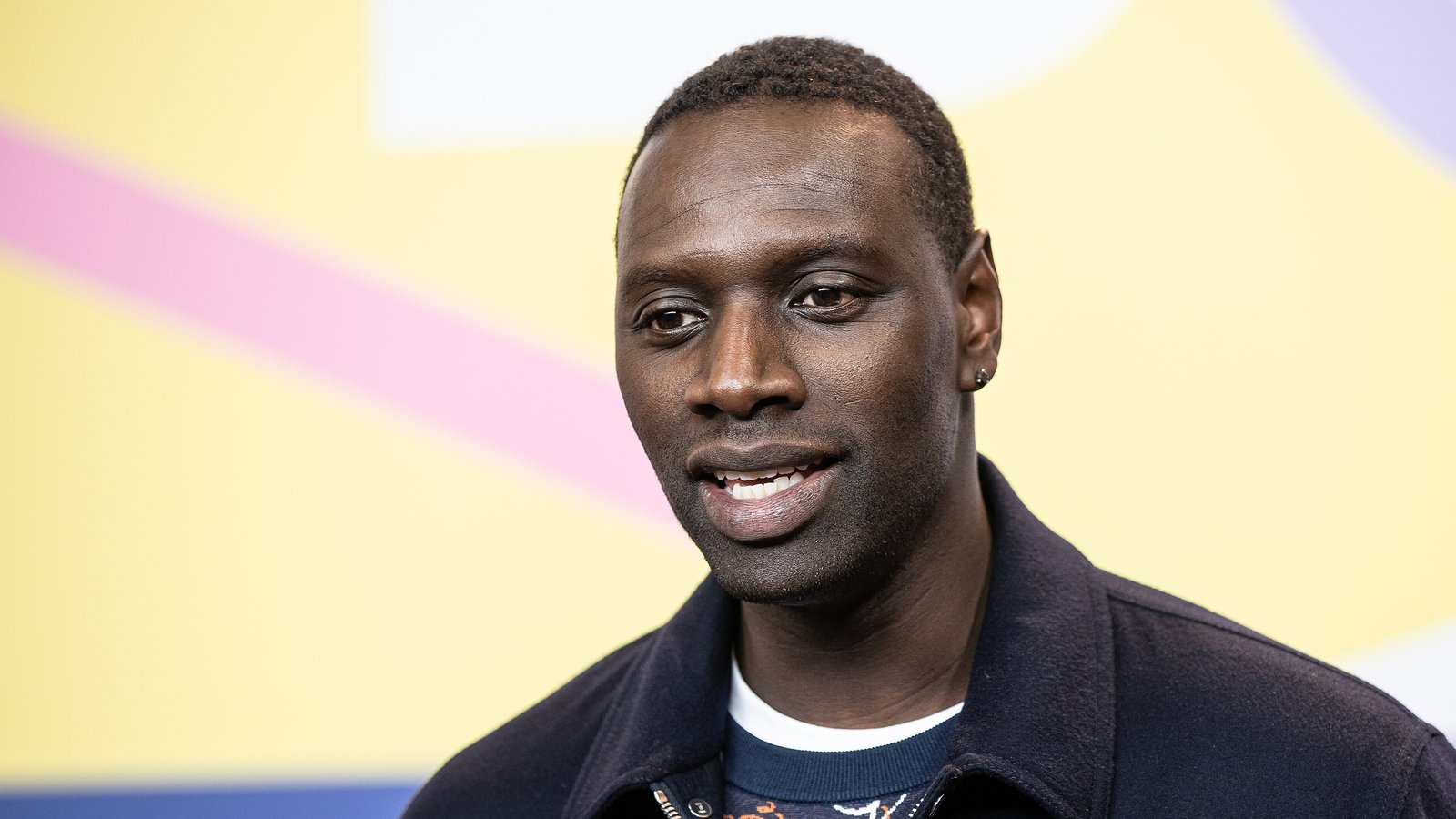 French actor Omar Sy, in a navy jacket, smiles in front of a bright yellow and pink backdrop at the Berlin International Film Festival.