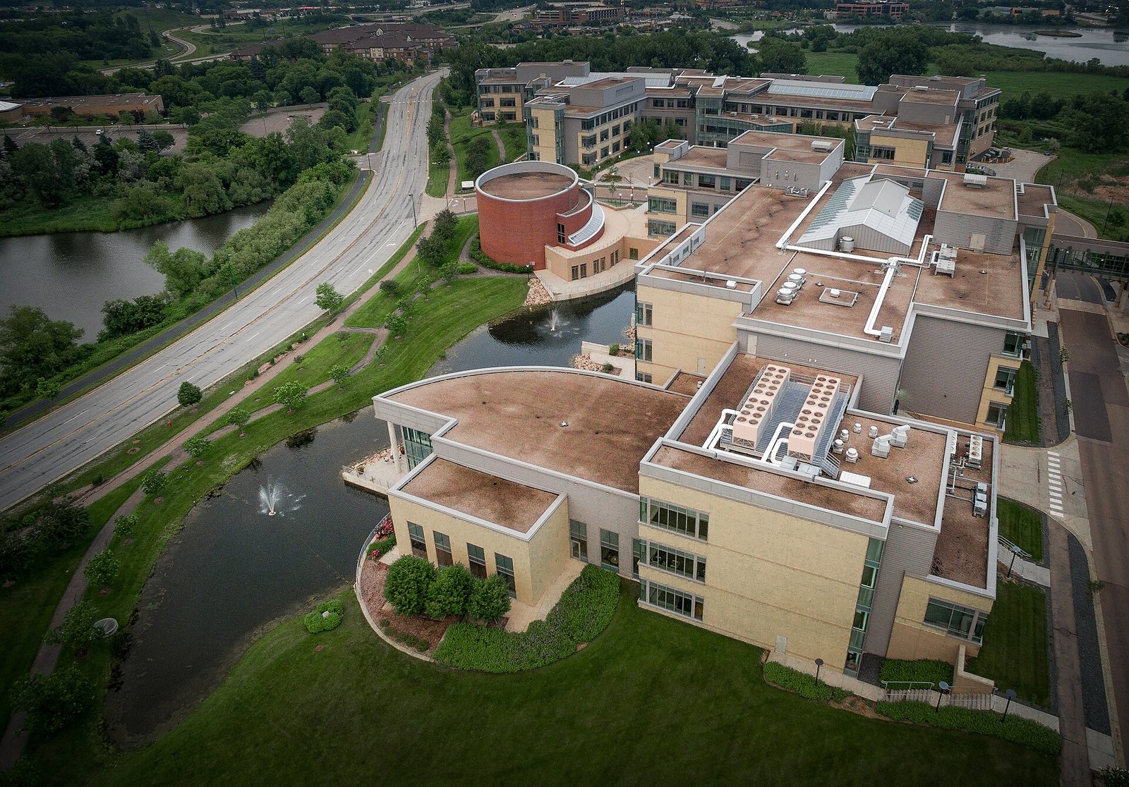 Aerial view of a large beige office campus with flat roofs, a round building, a pond and a curving road lined with green trees and lawns.