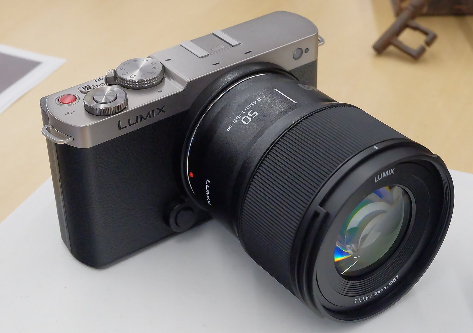 A silver and gray Panasonic Lumix S9 compact full-frame camera with a black Lumix 50mm prime lens, on a white table.