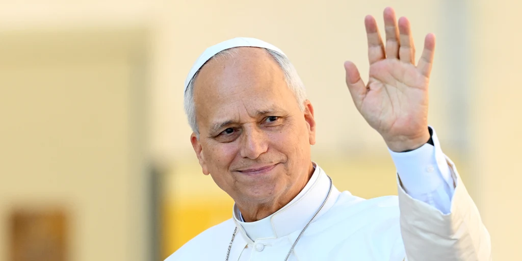 Pope Leo XIV in white cassock and zucchetto, smiling and waving with his left hand.