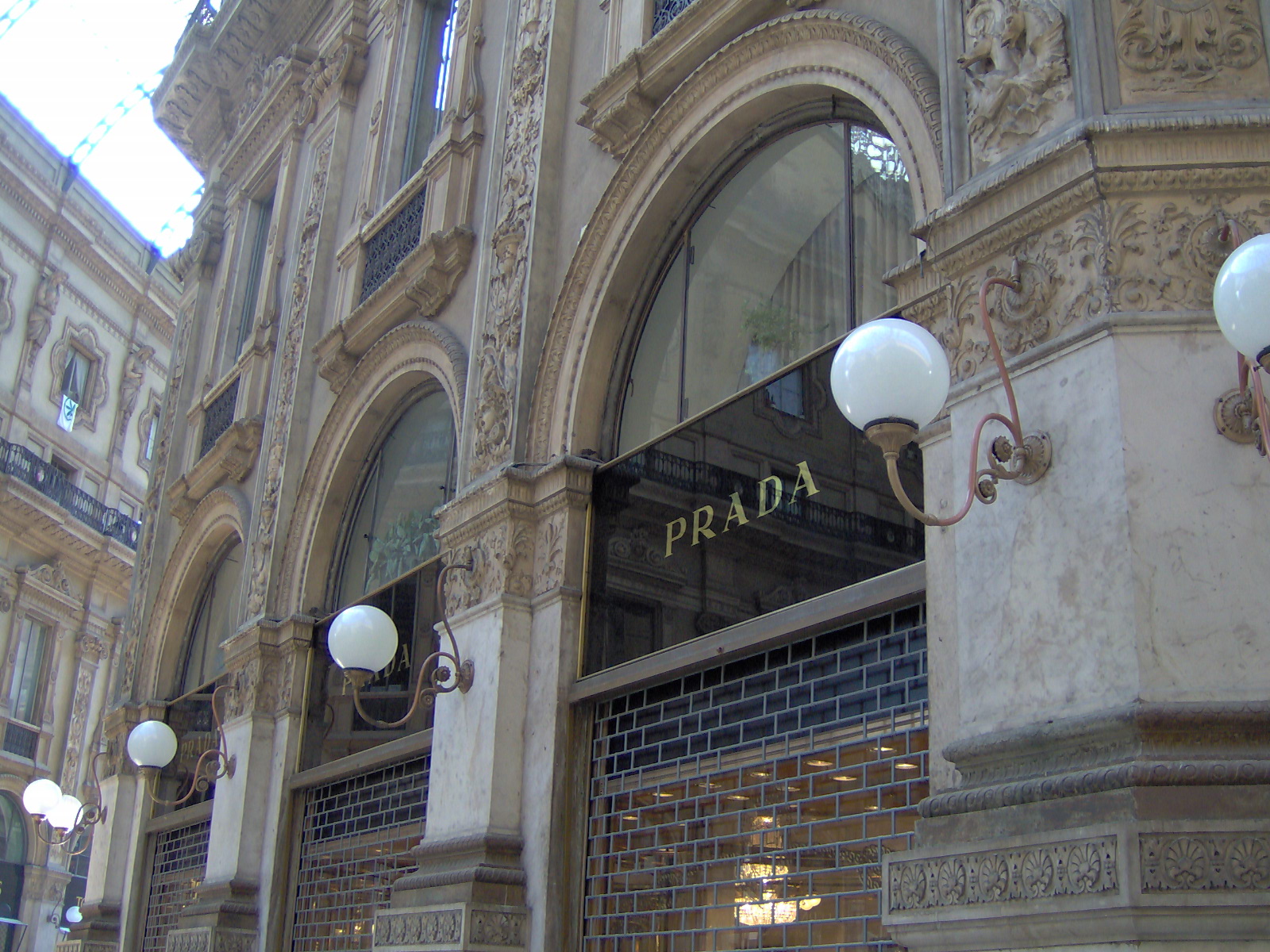 Ornate stone arches of the Galleria Vittorio Emanuele II in Milan, with gold PRADA lettering on black glass above the boutique's shuttered windows and globe lamps on curved brackets.