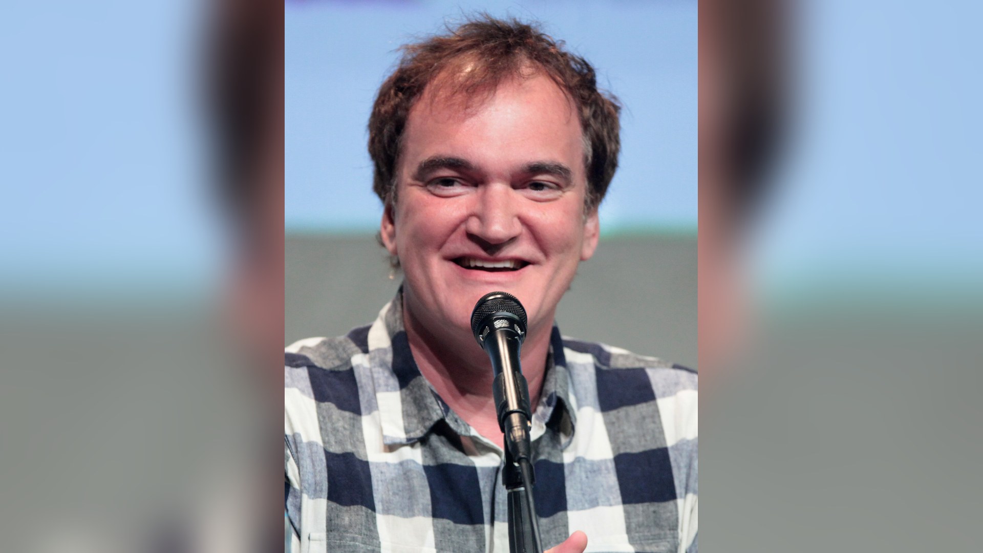 Quentin Tarantino, smiling, in a blue-and-white checked shirt, speaking into a microphone at a Comic-Con panel.