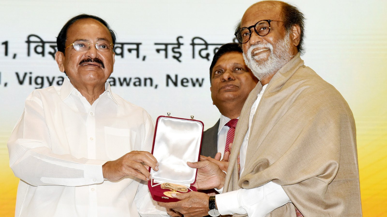 Rajinikanth, with a white beard and glasses and a beige shawl, smiles as he accepts the Dadasaheb Phalke Award medal on stage in New Delhi.