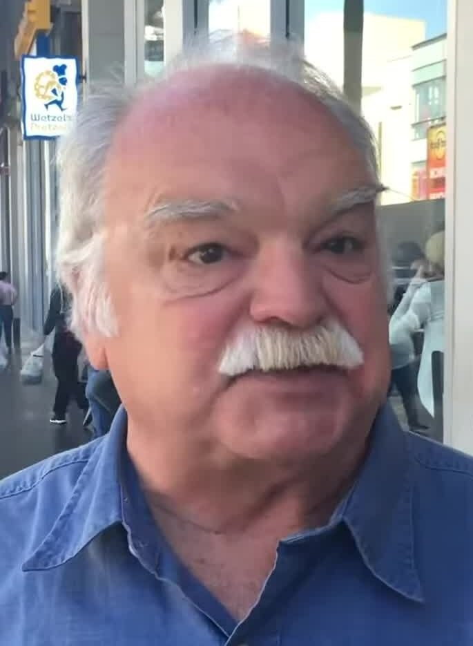 Richard Riehle, a white-haired actor with a gray mustache in a blue denim shirt, speaking on a Los Angeles street in 2016