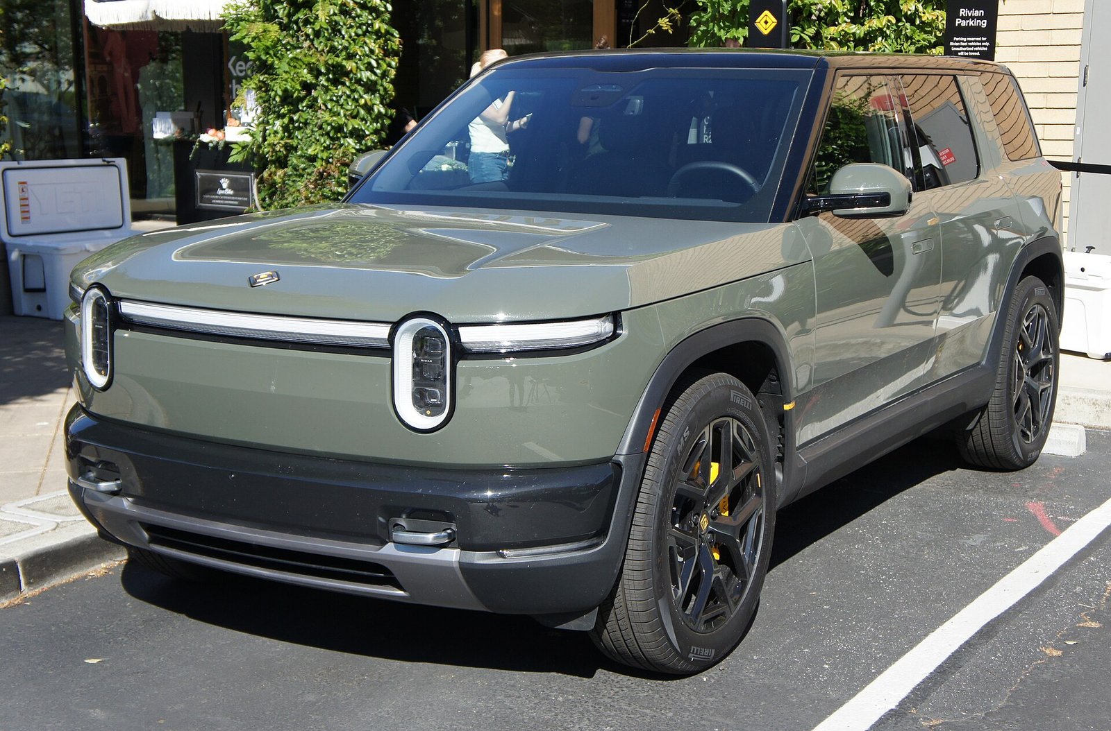 A sage green Rivian R2 electric SUV with oval headlights and a light bar, parked on a street.