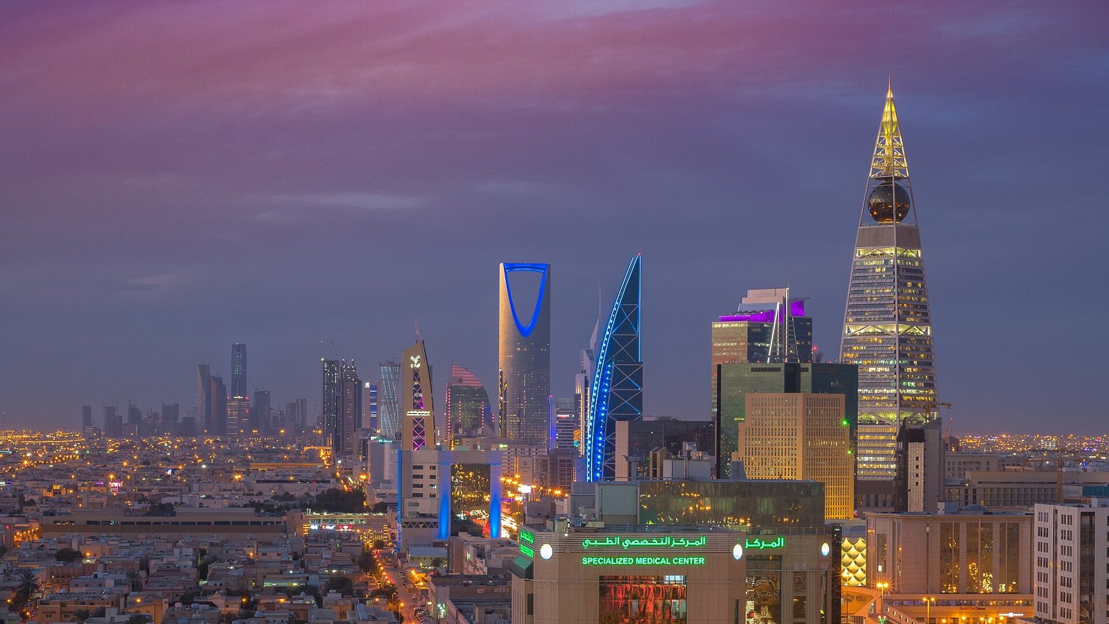The skyline of Riyadh at dusk, with the Kingdom Centre's arched tower lit in blue and other high-rises glowing against a purple sky.