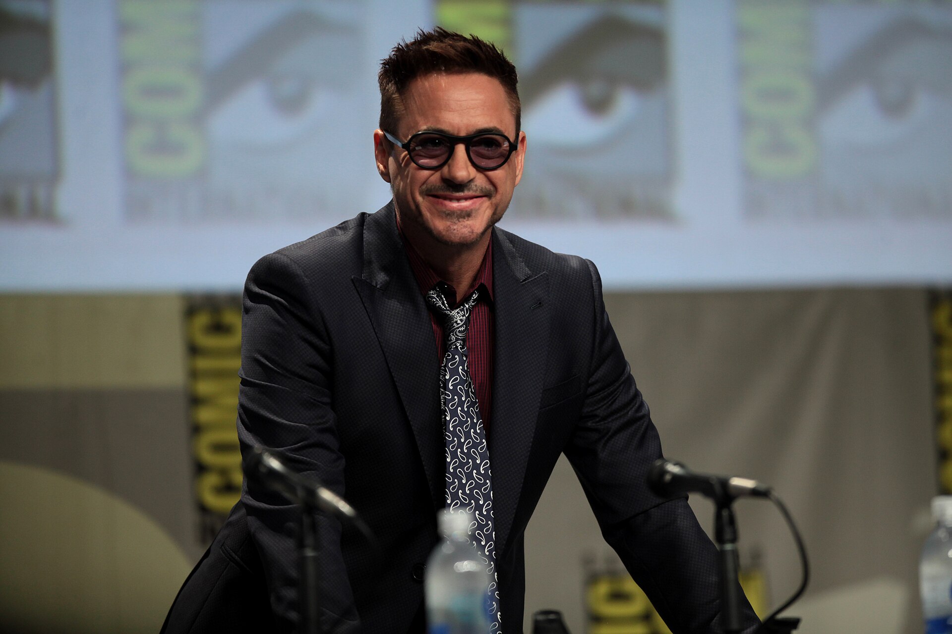Robert Downey Jr. in round dark glasses and a dark suit smiling at a microphone on a Comic-Con panel stage.