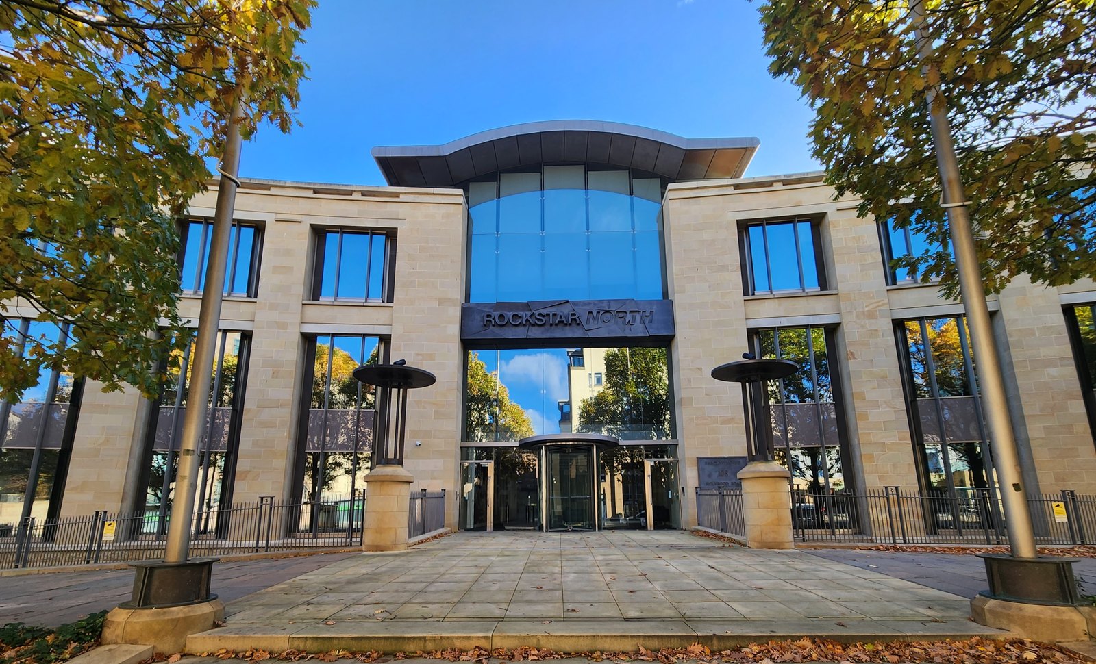 The stone and glass front entrance of the Rockstar North studio building in Edinburgh under a blue sky, with the Rockstar North sign above a revolving door.