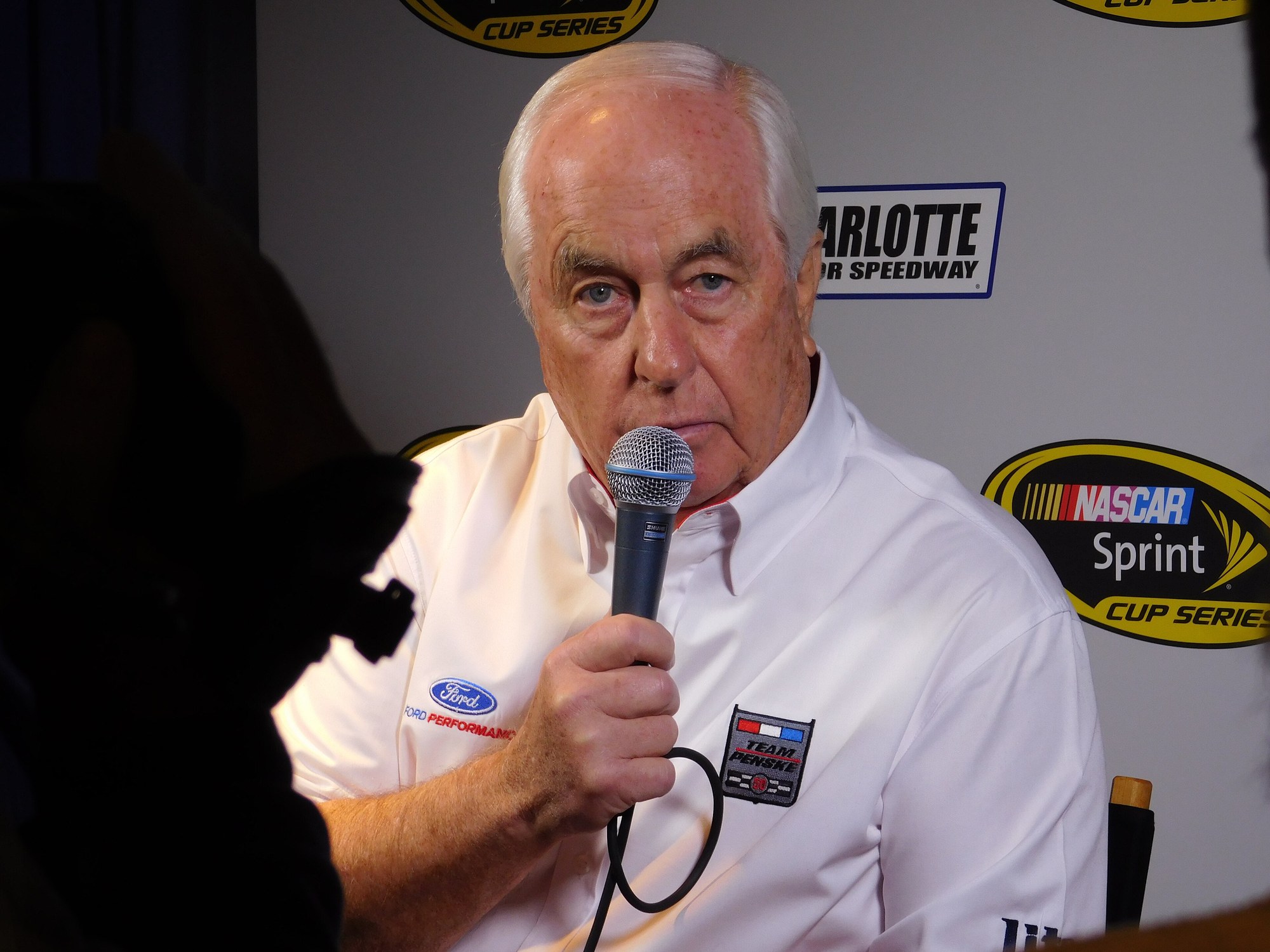 Roger Penske, white-haired and wearing a white Team Penske shirt, speaks into a handheld microphone in front of a NASCAR backdrop.