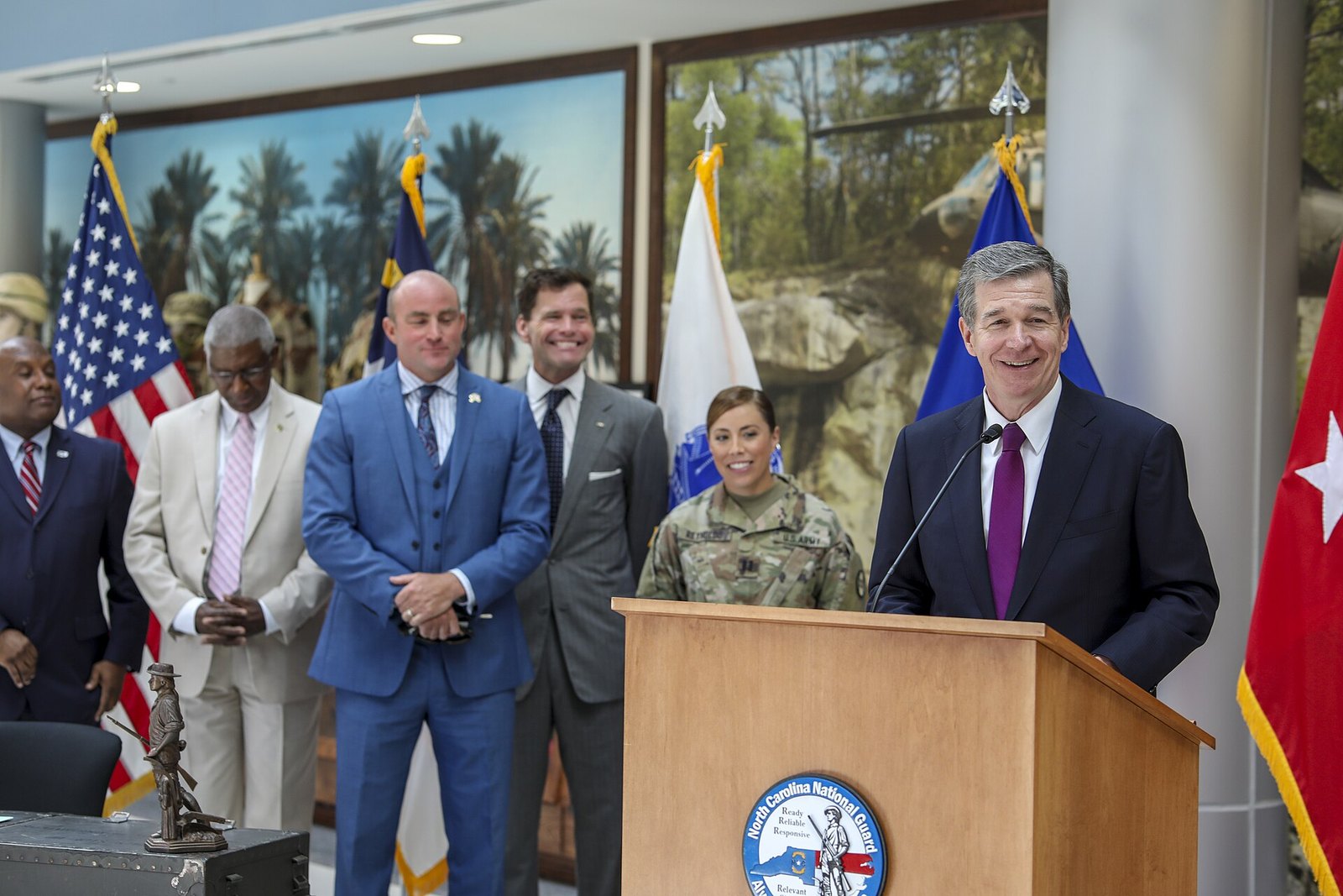 Roy Cooper, in a dark suit, speaks into a microphone at an outdoor bill-signing ceremony with uniformed North Carolina National Guard members behind him.