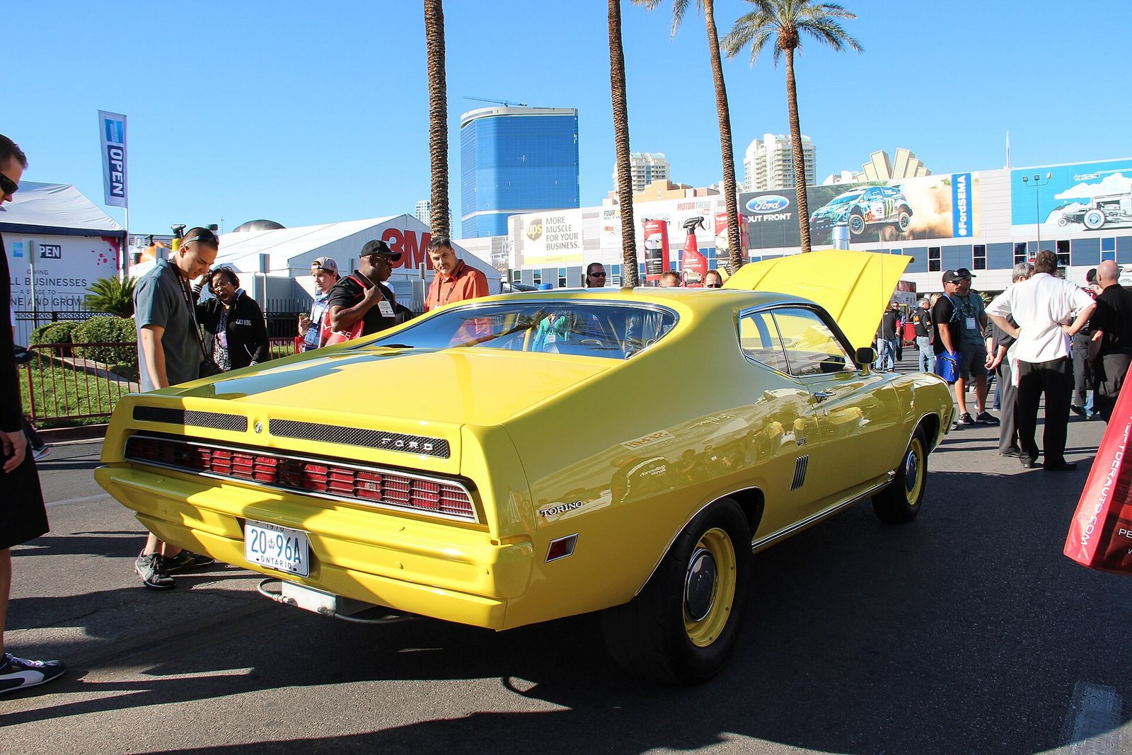 Crowds walking past a yellow classic muscle car with its hood up at an outdoor display at the SEMA Show in Las Vegas.