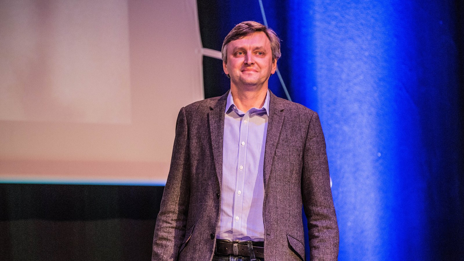 Film director Sergei Loznitsa, in a grey tweed jacket and striped shirt, smiles on a stage lit with blue light.