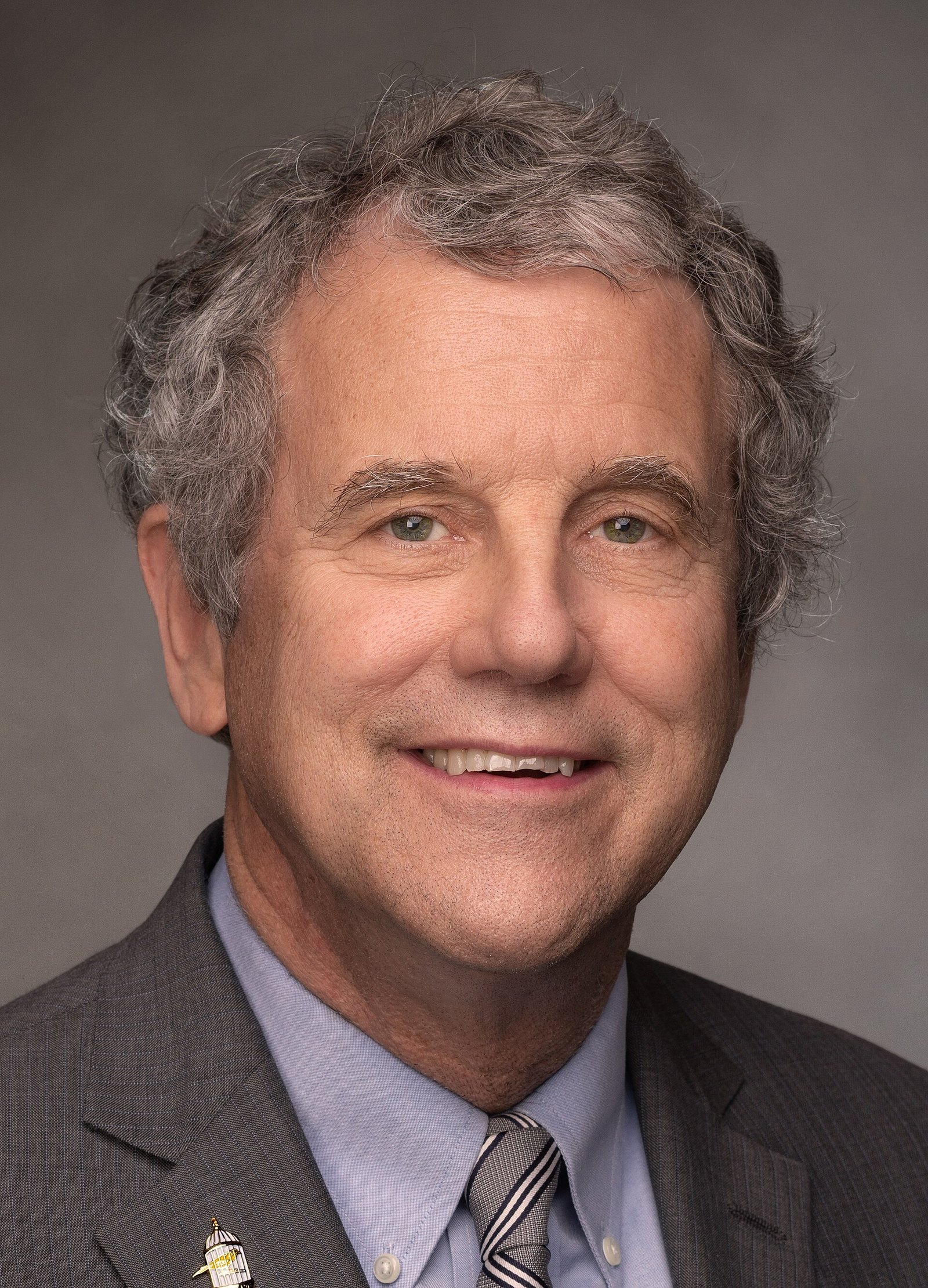Official portrait of Sherrod Brown, with curly gray hair, in a gray suit and striped tie, smiling against a dark backdrop.