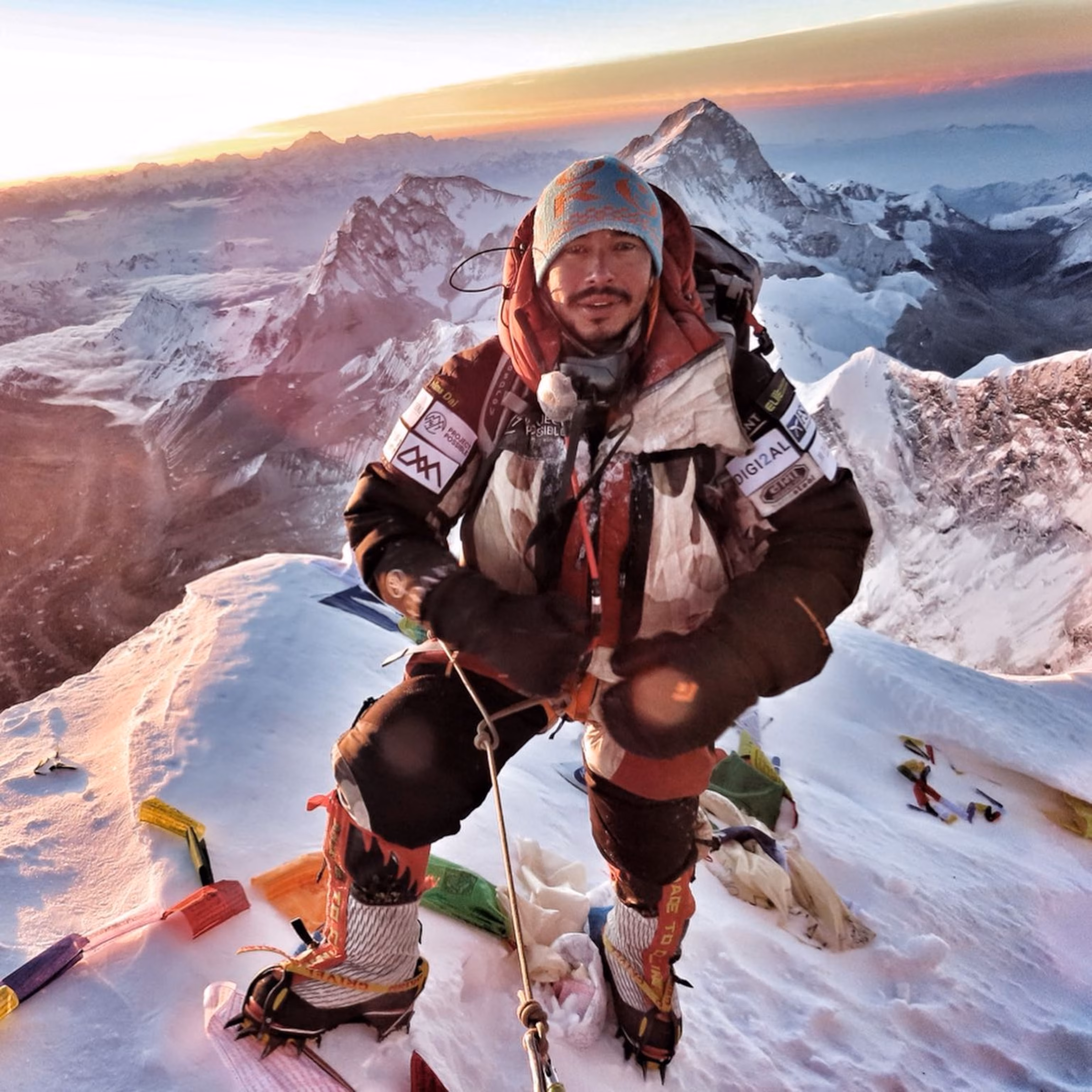 Nirmal Purja in expedition clothing on a snow-covered ridge with mountains behind him.
