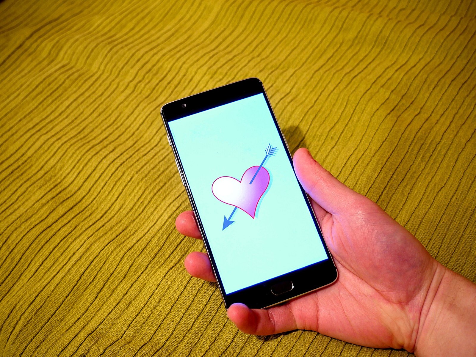 A hand holding a black smartphone against a yellow textured background, the screen showing a pink heart pierced by a blue arrow on a light blue background.