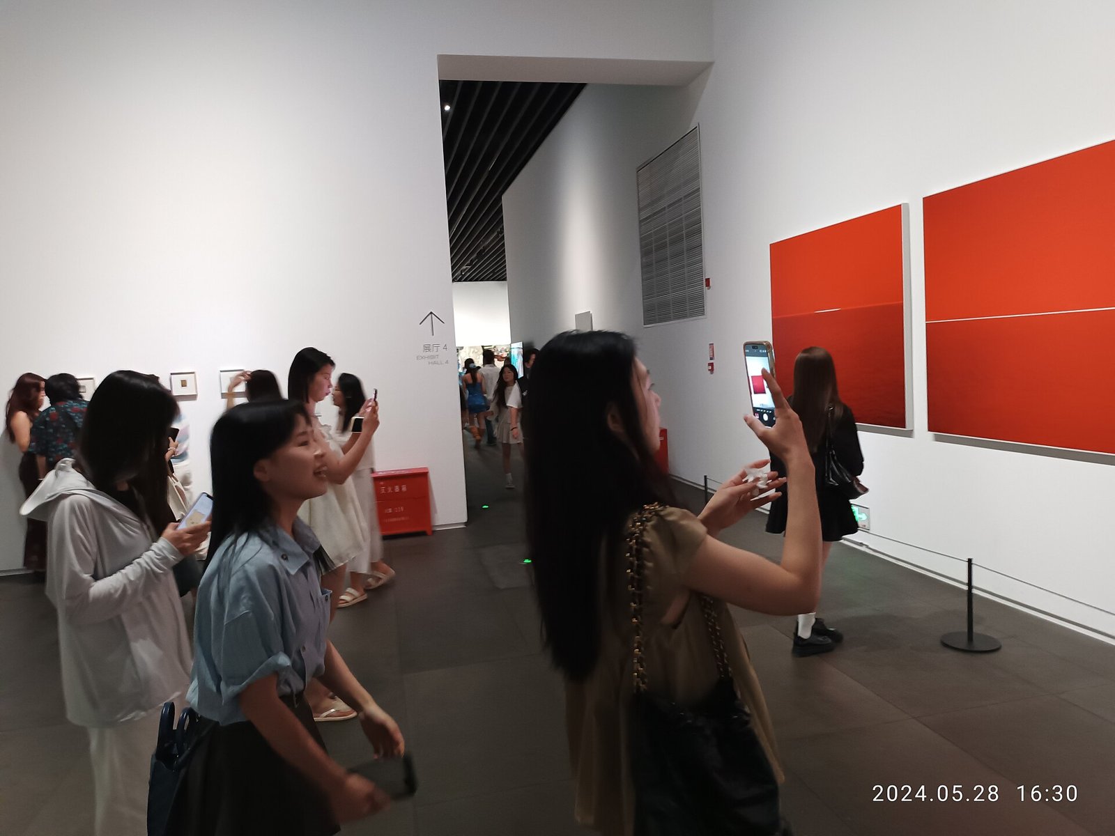 Several women holding up smartphones to photograph large red paintings on the white walls of an art museum gallery.
