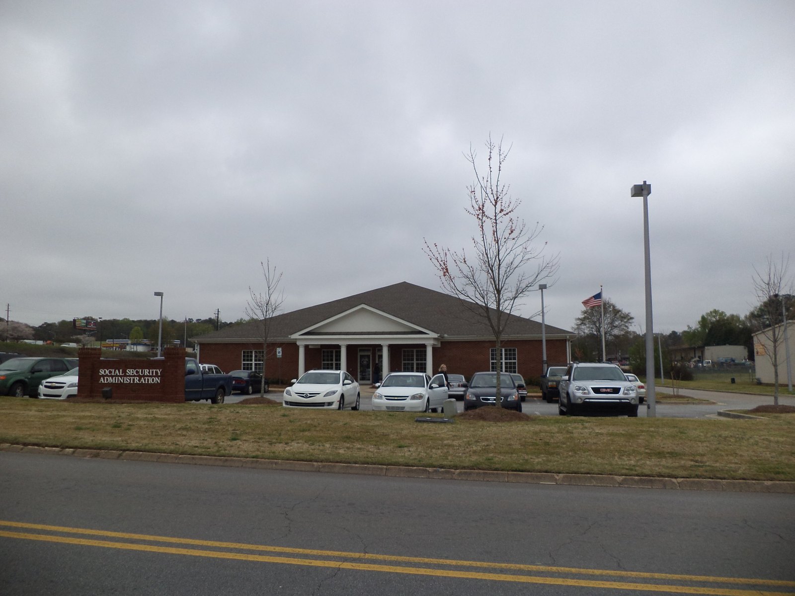 A single-storey brick Social Security Administration office with a white-columned entrance, cars in the parking lot and a sign reading Social Security Administration on the lawn under a grey sky.