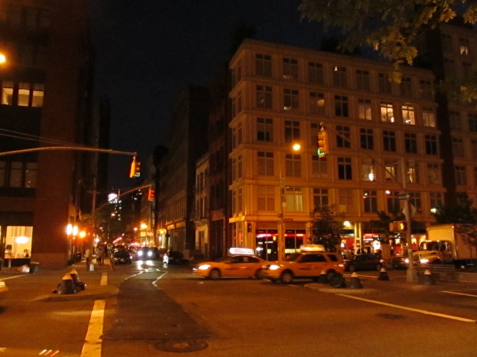 A SoHo street intersection in New York at night, with yellow taxis, streetlights and lit storefronts beneath tall brick and cast-iron buildings.