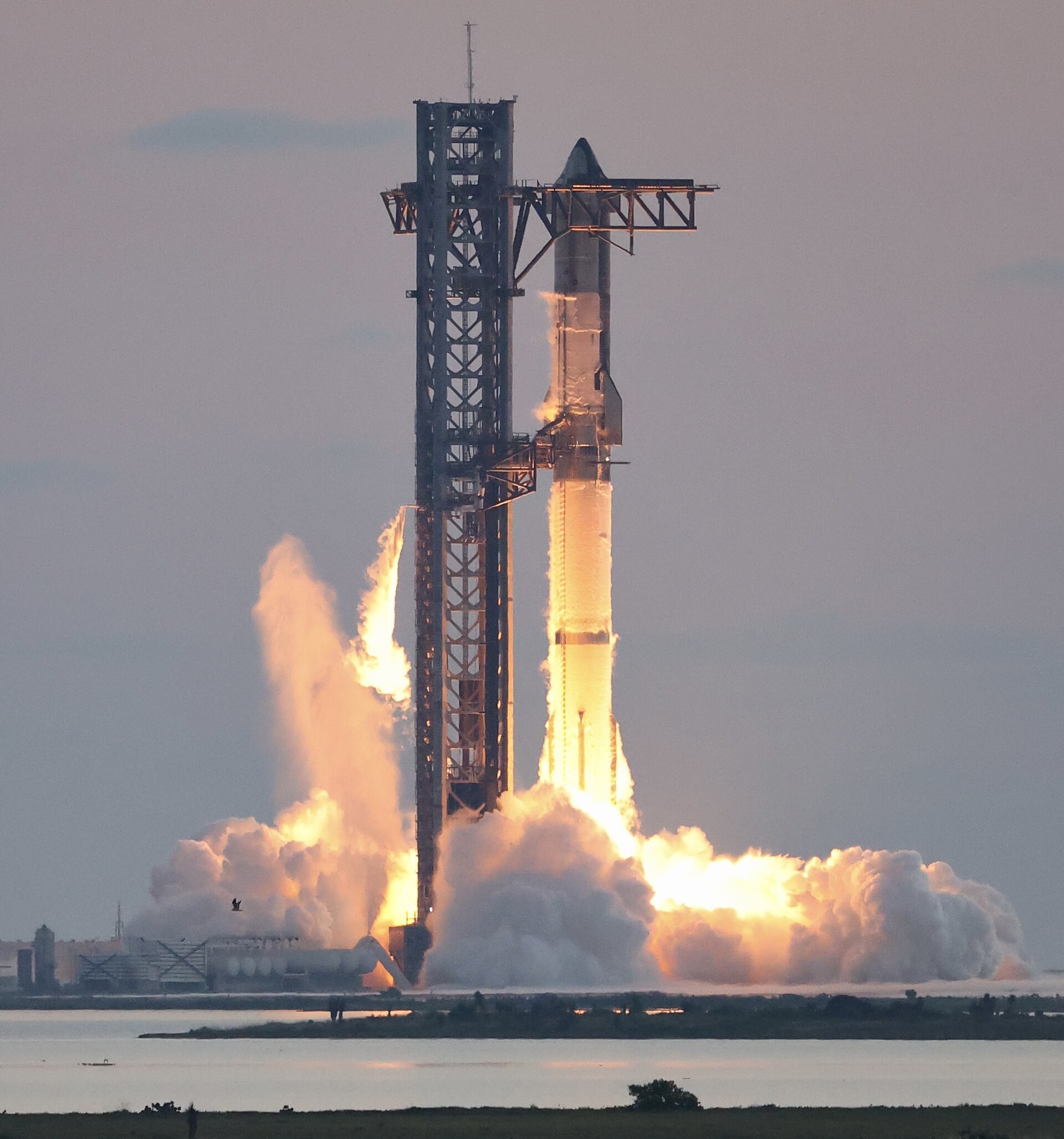 SpaceX's stainless-steel Starship rocket lifting off beside its launch tower in a cloud of fire and steam