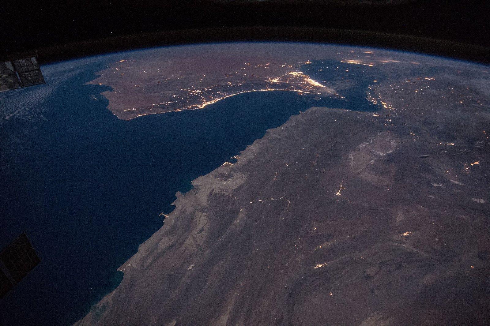 A night view from space of the Strait of Hormuz, with the dark mass of the Musandam peninsula and the lights of Gulf cities along the coast.