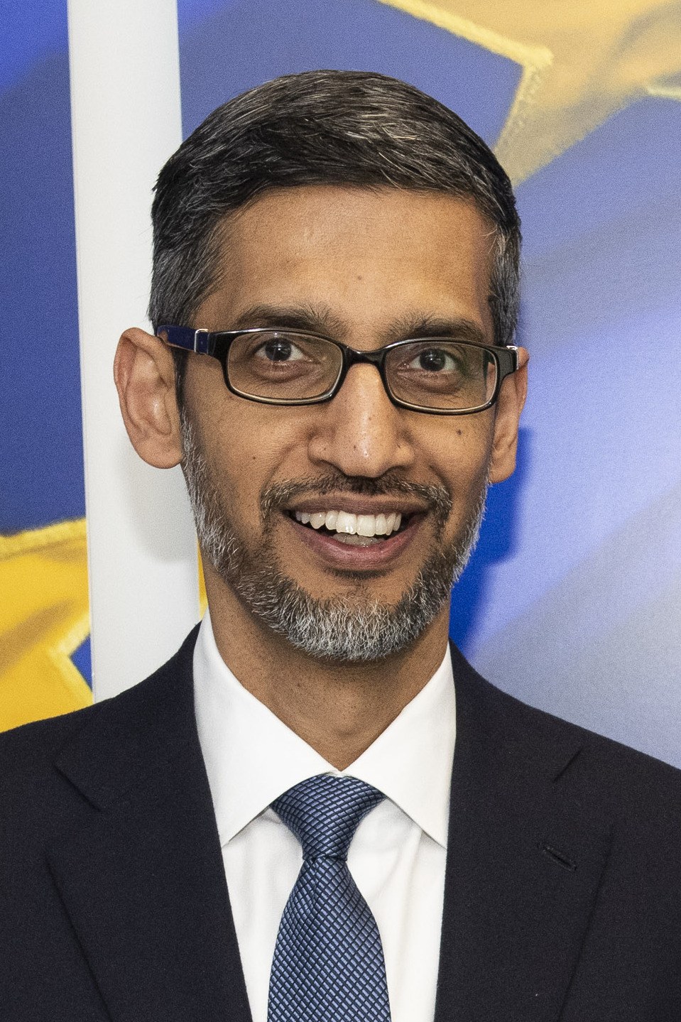 Portrait of Google CEO Sundar Pichai in a dark suit and white shirt, smiling, against a light background.