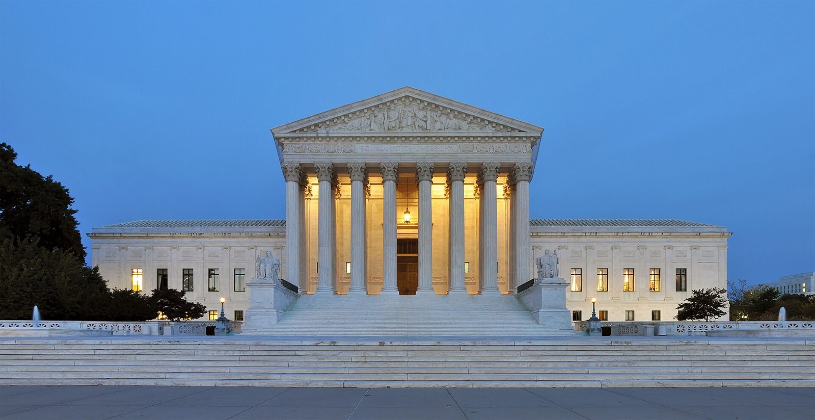 The white marble U.S. Supreme Court Building in Washington, D.C., lit from within at dusk, with its columned front and wide plaza under a blue sky.