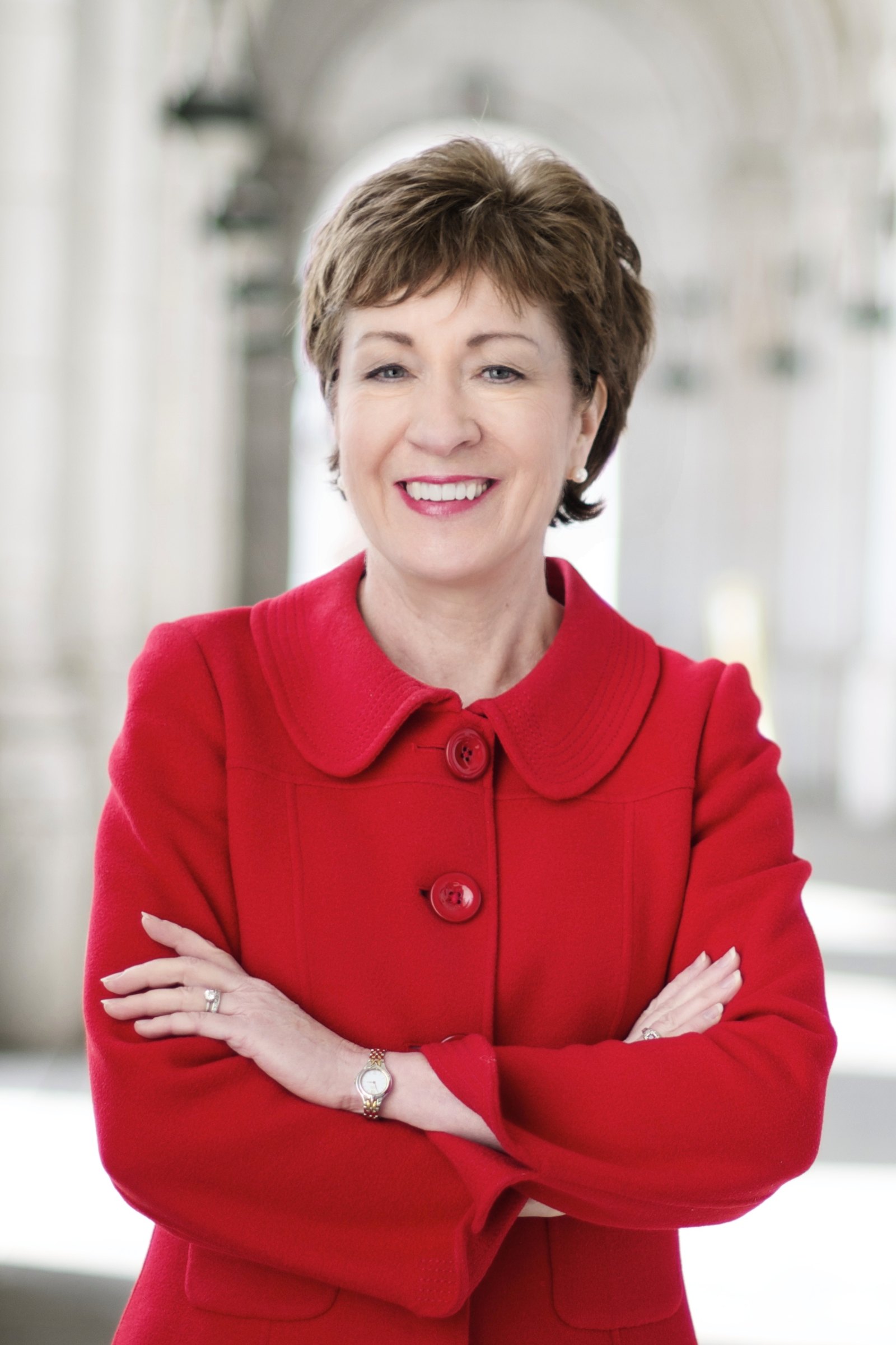 Official portrait of Senator Susan Collins in a red jacket, arms crossed and smiling, against a soft white background.
