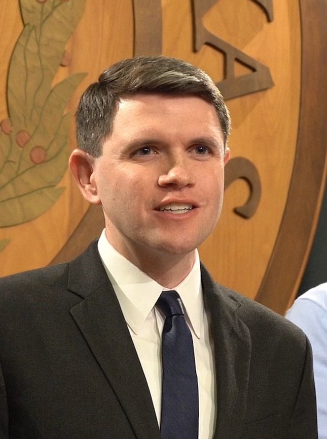 James Talarico, in a dark suit and tie, smiles at a press conference inside the Texas Capitol in Austin, with an ornate seal behind him.