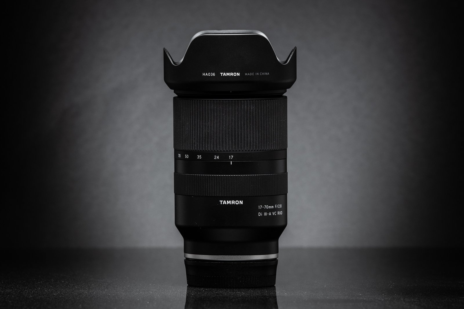 A black Tamron 17-70mm f/2.8 zoom lens with its petal-shaped hood attached, standing upright against a dark gray background.