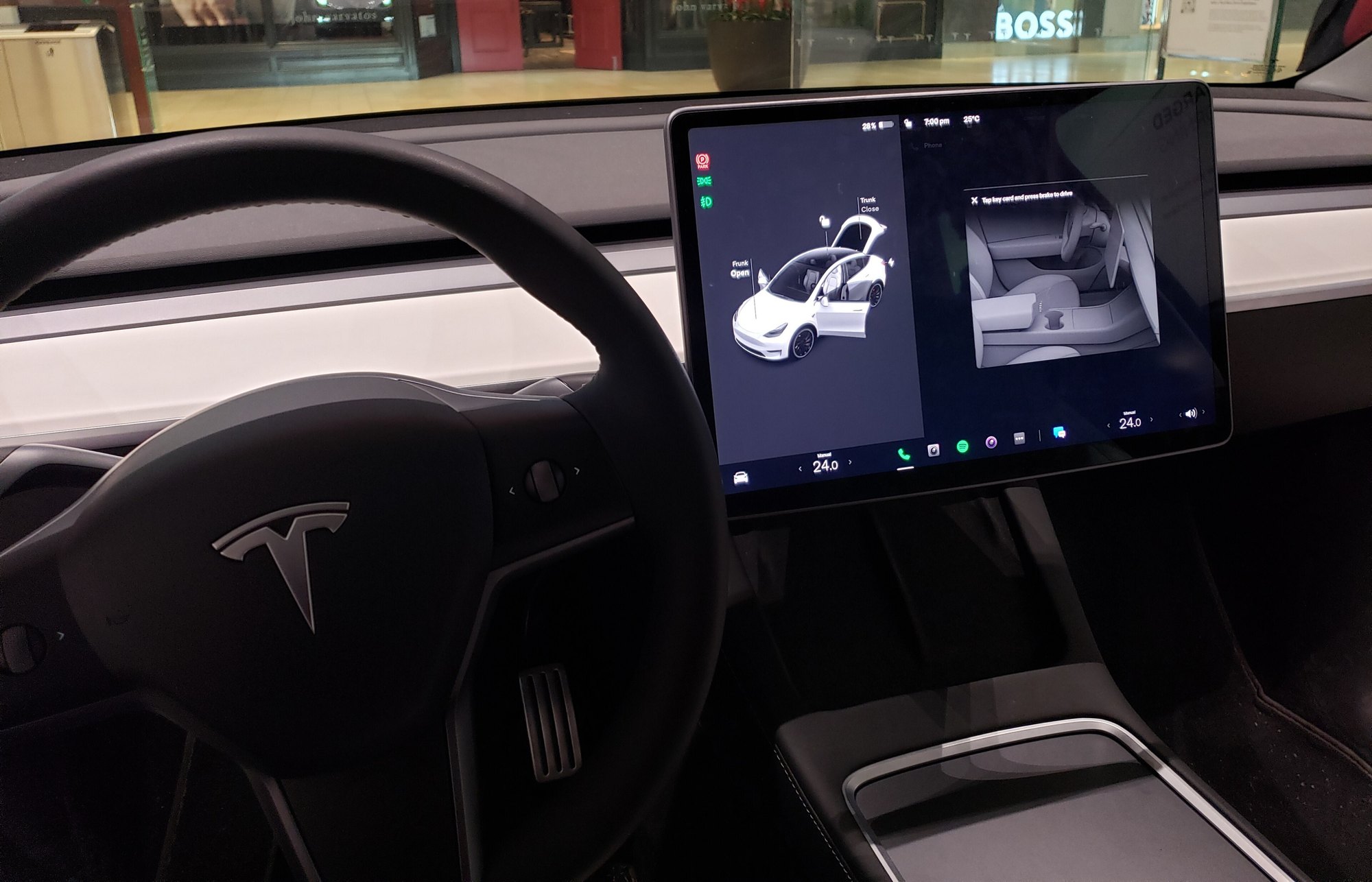 Inside a Tesla Model 3: a black steering wheel with the Tesla logo and a large central touchscreen showing a 3D image of the car with its trunk and frunk open.