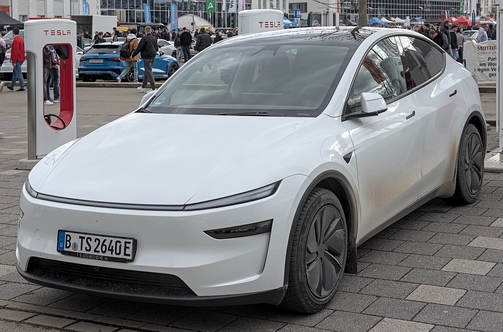 A white redesigned Tesla Model Y electric SUV with a full-width light bar, parked near Tesla Superchargers.