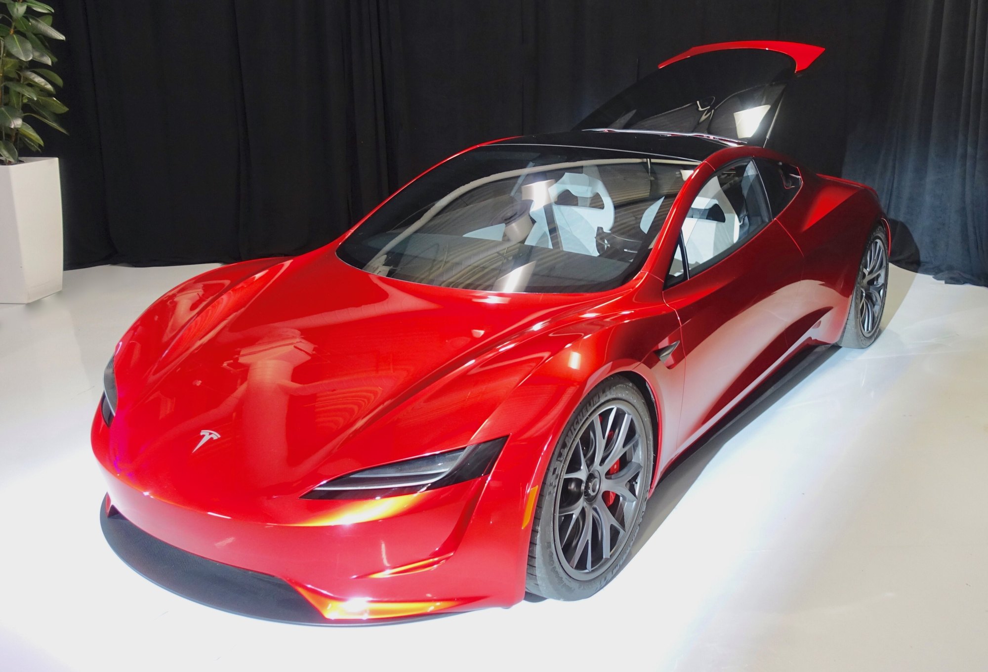 A glossy red Tesla Roadster prototype with a glass roof and silver wheels displayed indoors against a black curtain.