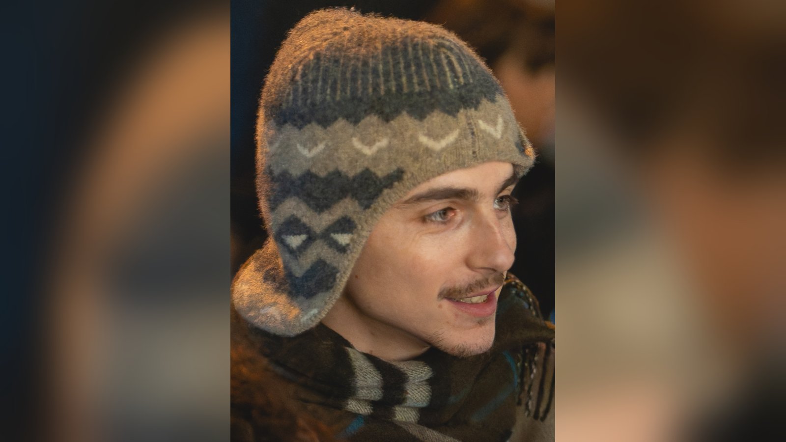 Timothée Chalamet, wearing a patterned knit hat with ear flaps and a striped scarf, smiles at an evening event in London.