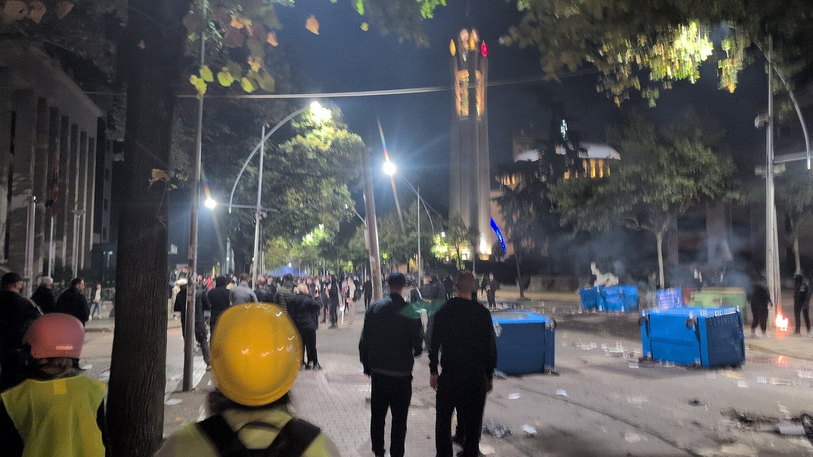 Protesters, some in hard hats, walk down a littered street in Tirana at night under streetlights, with overturned bins and smoke in the background.