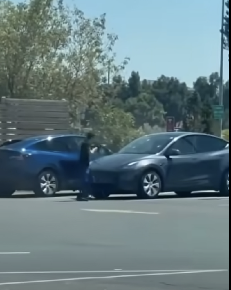 A distant, low-resolution frame showing a person standing beside a blue car in a car park, with a second dark car in the foreground.
