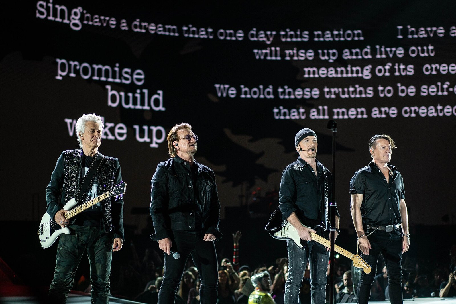 The four members of U2 stand in a line at the front of a stadium stage in black clothes, with lines of text projected on a giant screen behind them.