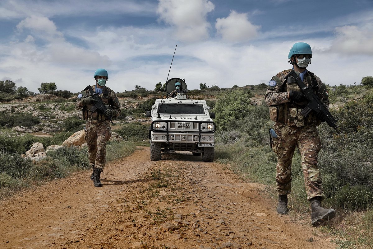 Two Italian soldiers wearing blue UN helmets and face masks walk along a dirt track in southern Lebanon, followed by a white armoured vehicle.