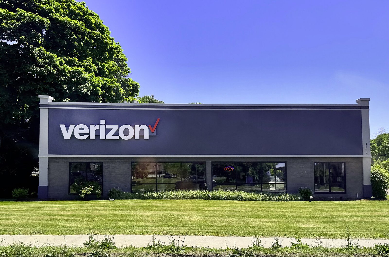 The front of a single-story Verizon retail store with a large white Verizon logo on a dark facade, behind a green lawn under blue sky.