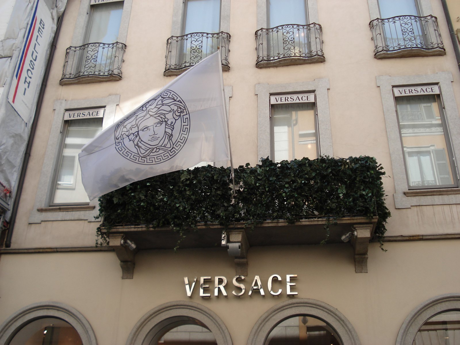 The cream stone facade of the Versace boutique on Via Montenapoleone in Milan, with a white Medusa-logo flag, a hedge-lined balcony and Versace lettering above the entrance.