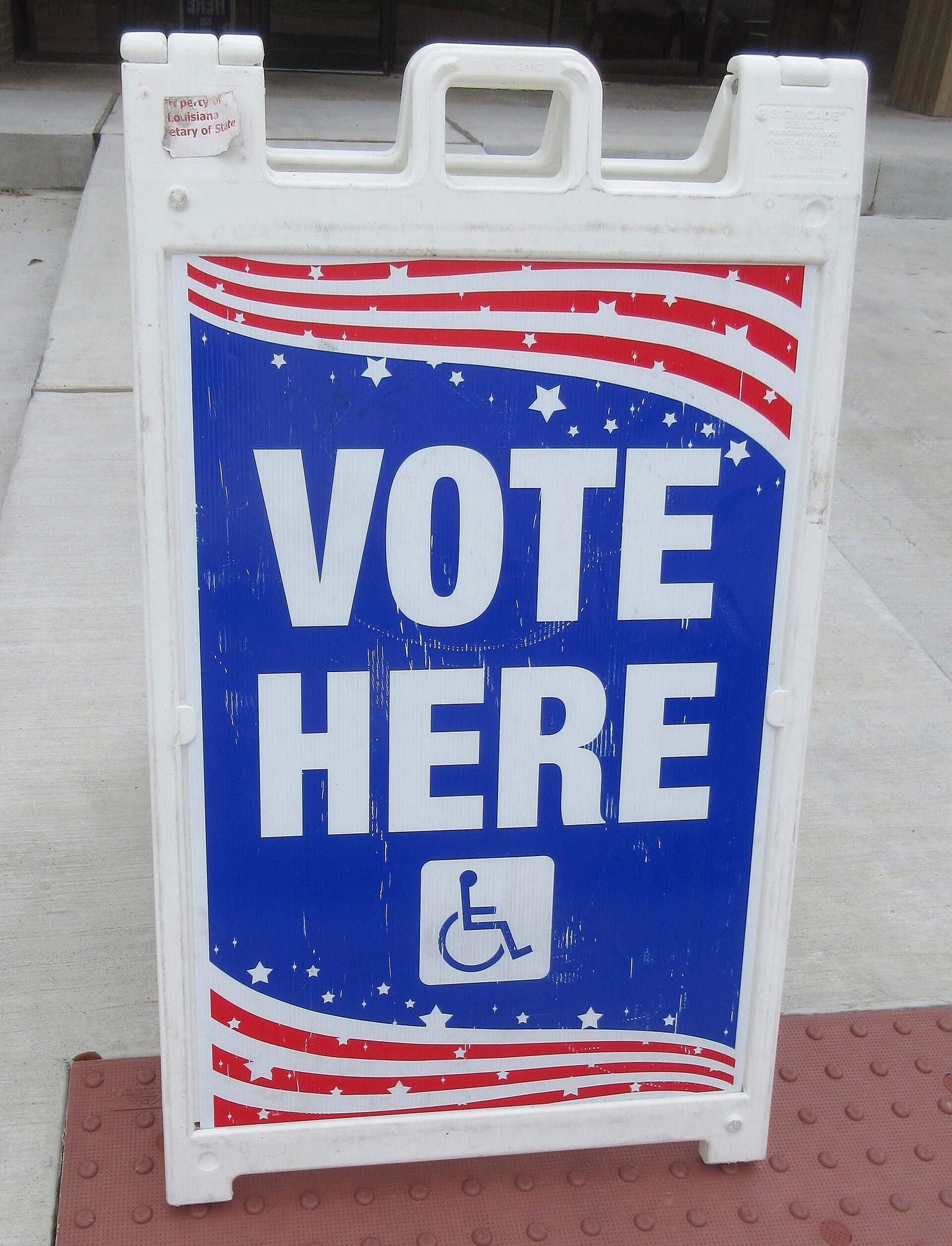 A white sandwich-board sign reading VOTE HERE in large blue letters with red stripes and a wheelchair-access symbol, standing on a sidewalk outside a polling place.