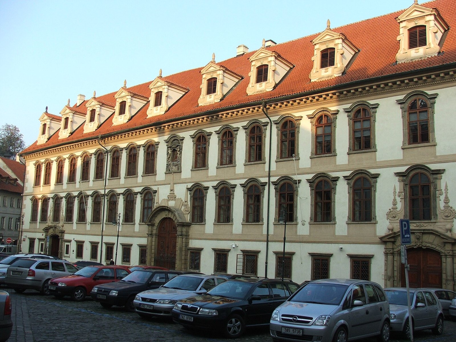 The pale yellow baroque facade of Wallenstein Palace in Prague, with a red-tiled roof and dormer windows, and cars parked in the square in front.