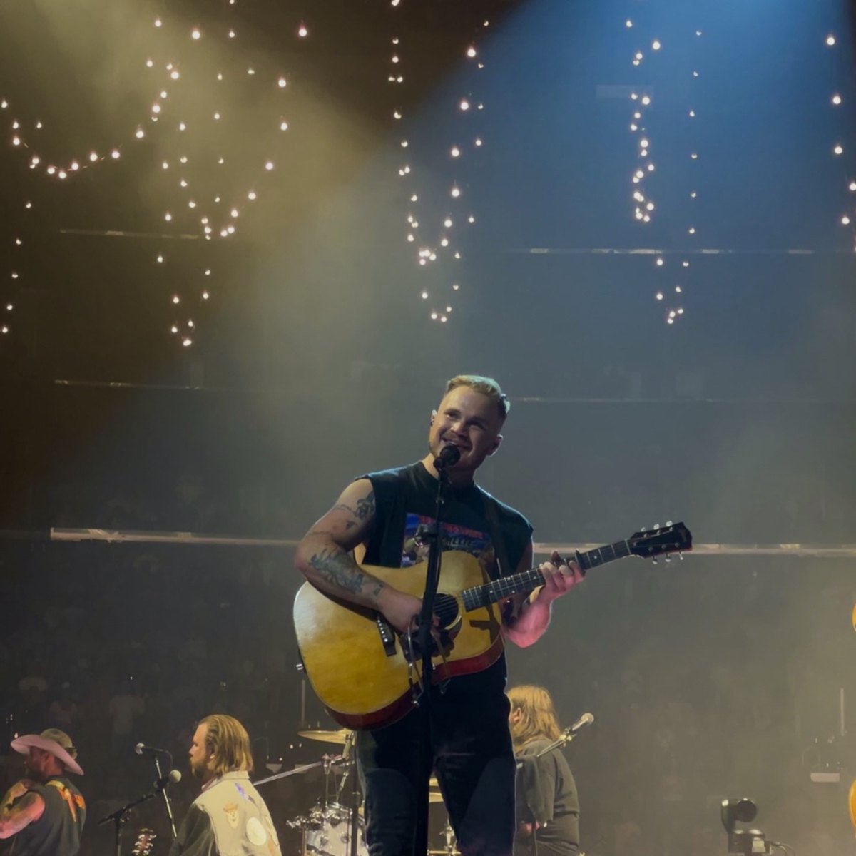 Zach Bryan sings and plays acoustic guitar on an arena stage under warm lights.